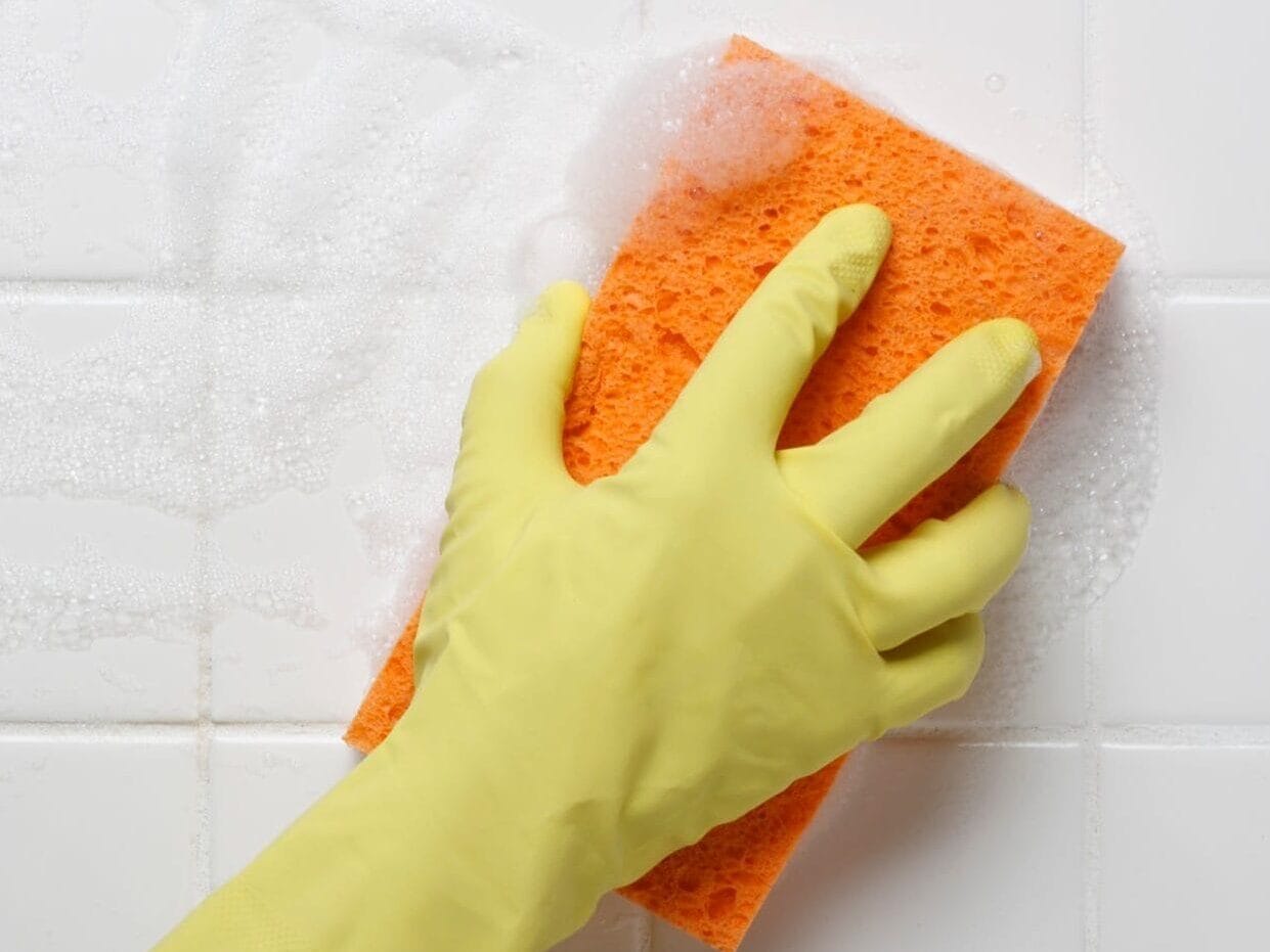 Close-up van een hand met een gele schoonmaakhandschoen die een oranje spons gebruikt om glanzende, witte badkamertegels te reinigen.